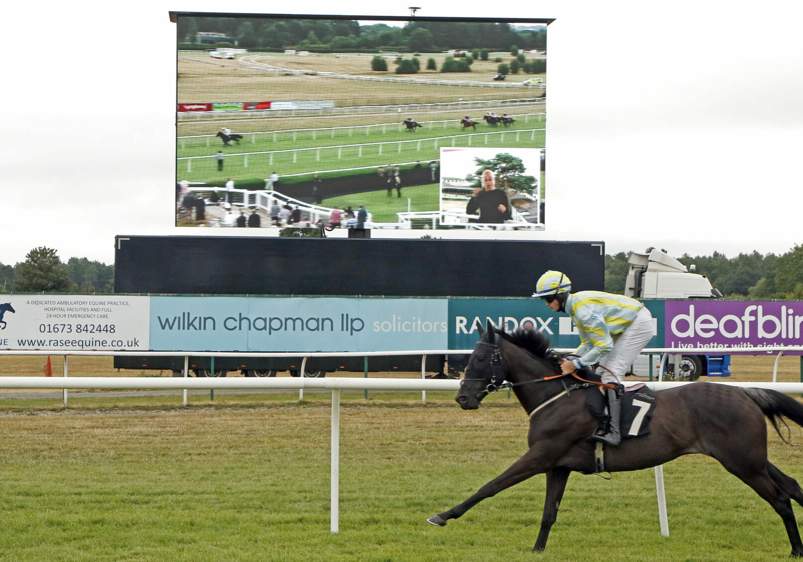 Racehorse and jockey galloping on a racecourse, with a large screen in the background featuring commentary in BSL.